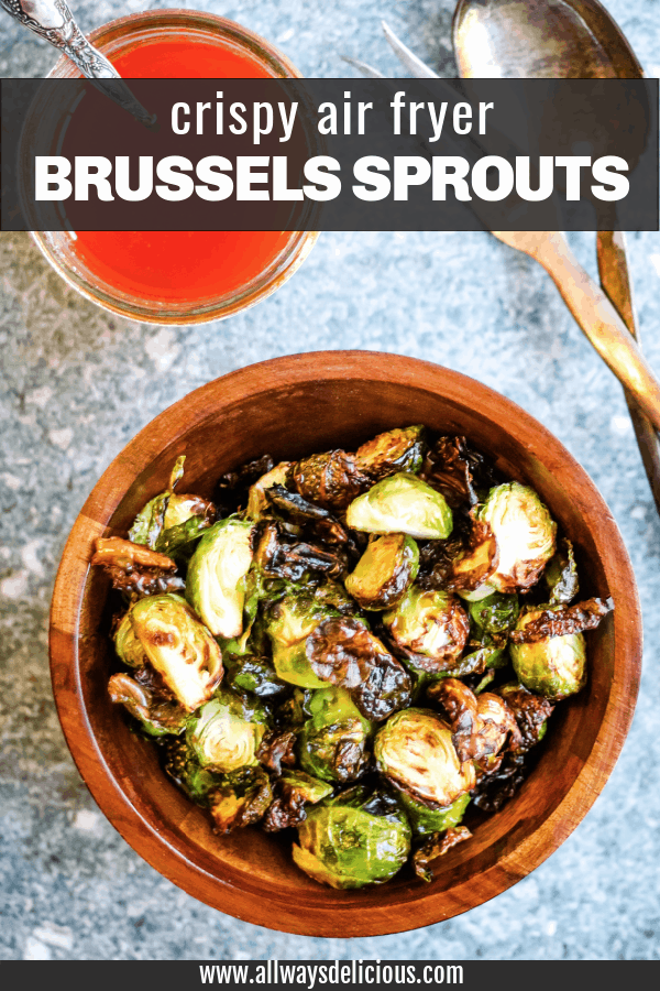Pinterest pin for air fryer fried brussels sprouts. Text says crispy air fryer brussels sprouts. image is an overhead shot of browned brussels sprouts in a wooden bowl on a stone counter top. there is a jar of harissa dressing and a pair of silver salad serves on the table next to the bowl.