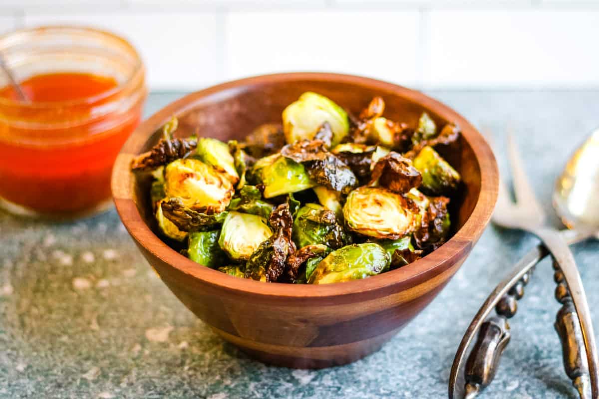 Air fried brussels sprouts, halved, browned, and crisp, in a wooden bowl. There is a jar of harissa dressing behind and to the left and a pair of silver salad servers on the table to the right.
