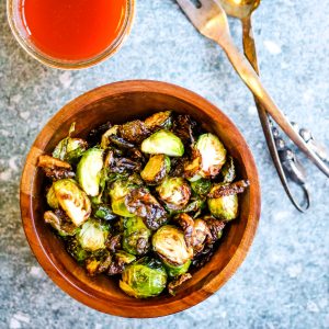 overhead shot of air fryer fried brussels sprouts in a wooden bowl with a jar of harissa dressing and a pair of silver salad servers on the side.