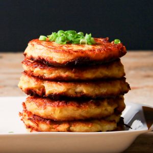 Low angle photo of a stack of 5 potato latkes on a white rectangular plate. There is a bowl of sour cream on the plate and both the sour cream and potato latkes are garnished with sliced green onions.