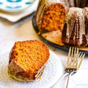 Low angle shot of a slice of pumpkin spice bundt cakeon a plate. There is the full cake wiht a piece taken out of it behind on a platter and 2 forks next to the serving plate.