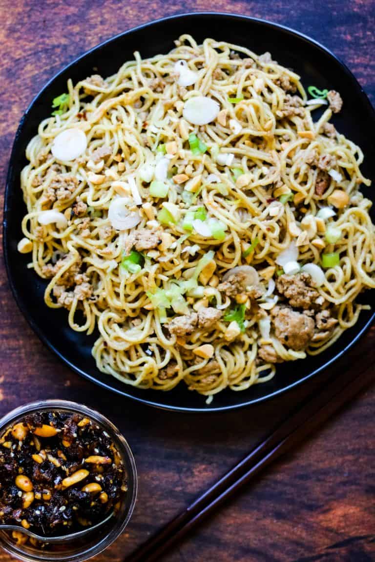 overhead shot of chinese sesame noodles on a black plate. Next to it is a blue and white serving bowl with sesame noodles in it and a pair of wooden chopsticks resting on the edge. There is also a jar of chile crisp. The table is dark brown wood.