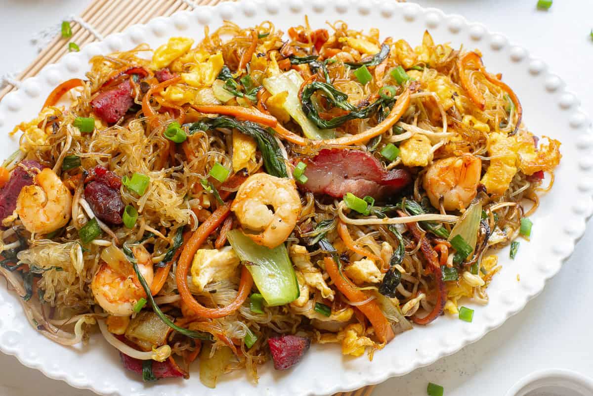 A plate of stir-fried singapore rice noodles with shrimp, BBQ pork, vegetables, egg, and green onions, served on a white platter.