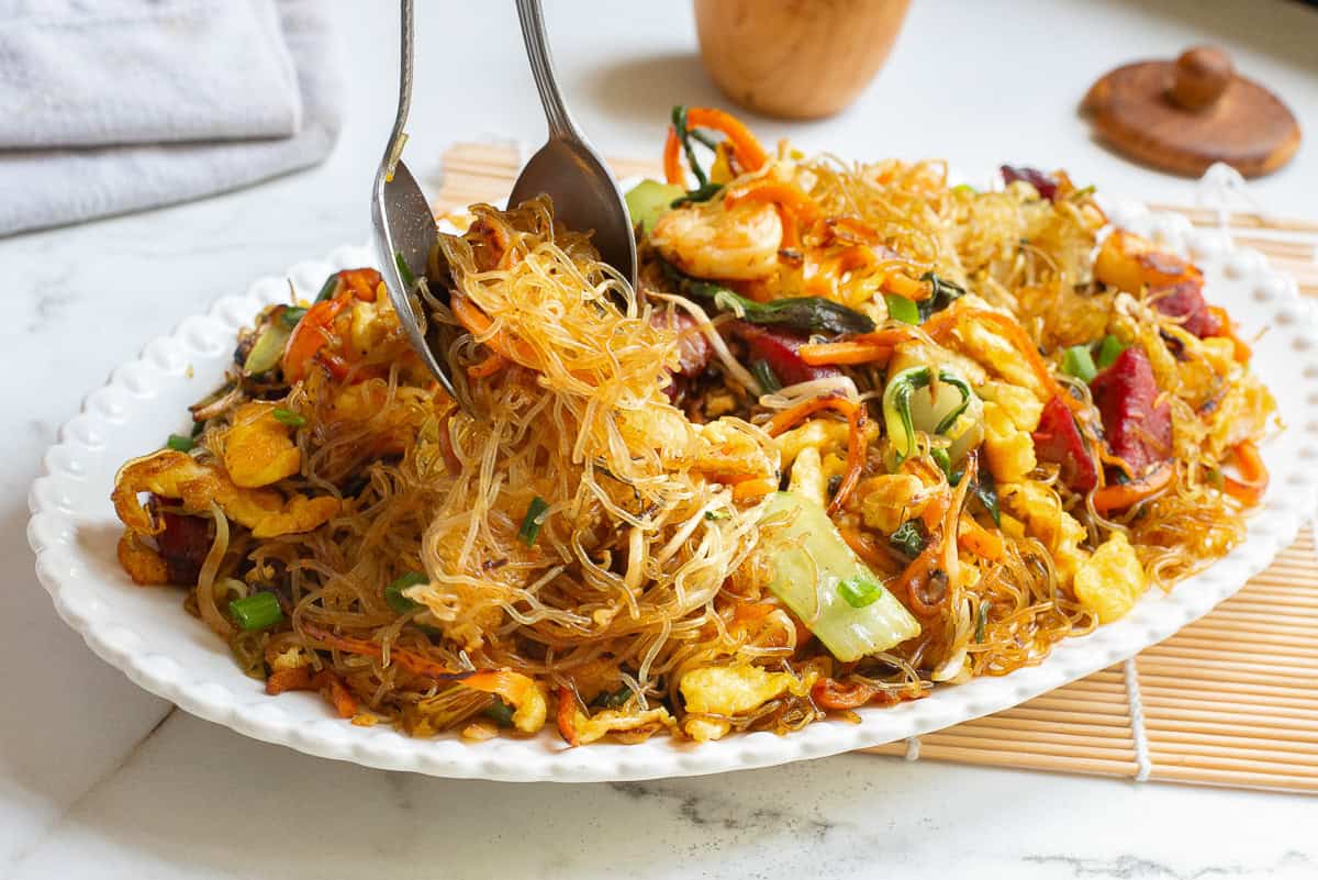 A plate of stir-fried singapore rice noodles with vegetables, egg, and meat pieces, served with metal tongs on a white dish.
