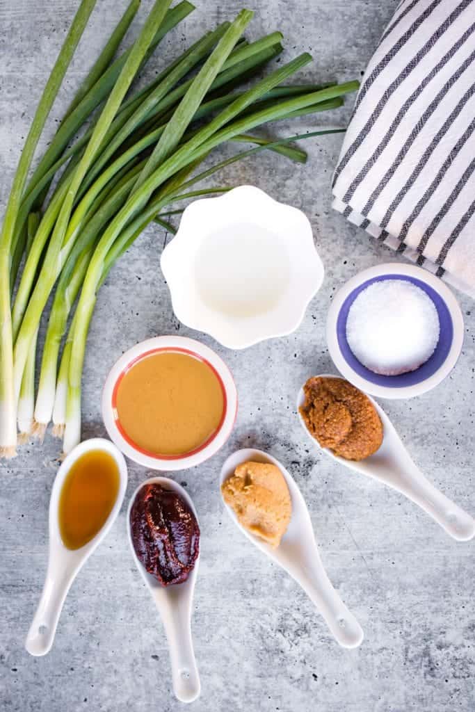 overhead shot of ingredients for miso ramen tare: sesame oil, goghujang, white miso paste, red miso paste, salt, rice vinegar, and sesame paste. There is also a bundle of green onions on the table next to the ingredients.