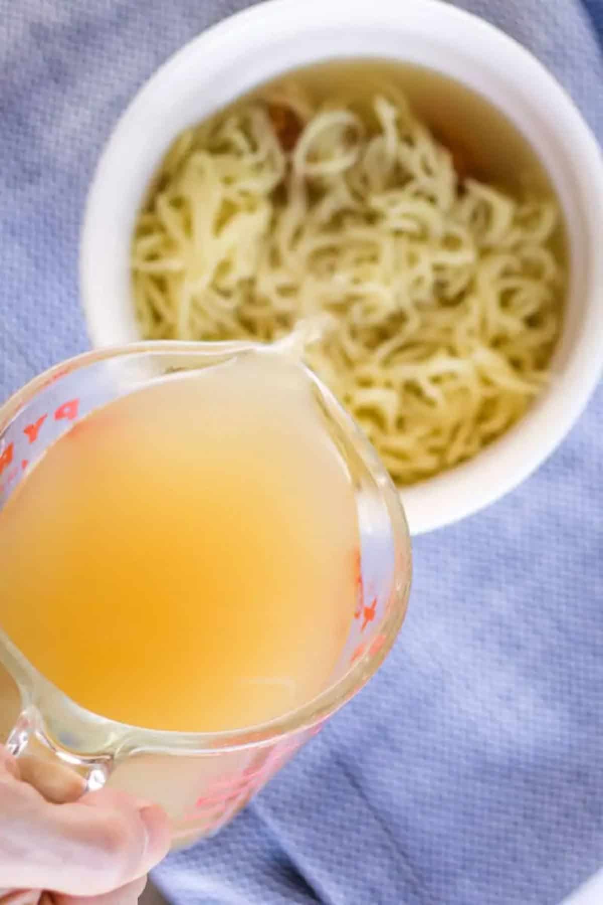 A hand pours light-colored broth from a measuring cup into a bowl of cooked miso ramen noodles on a blue textured cloth.