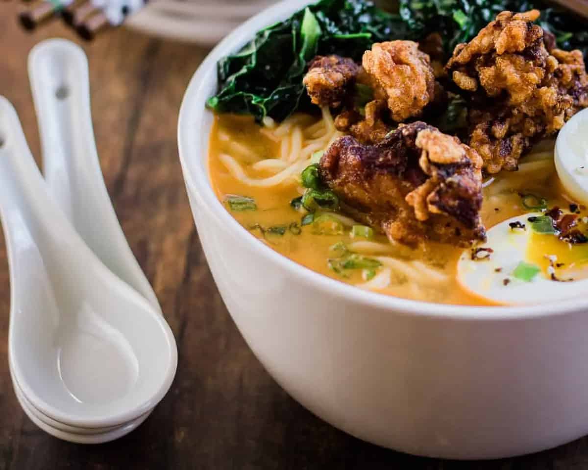 A bowl of miso ramen with fried chicken, boiled egg, greens, and broth sits on a wooden table next to two white ceramic soup spoons.