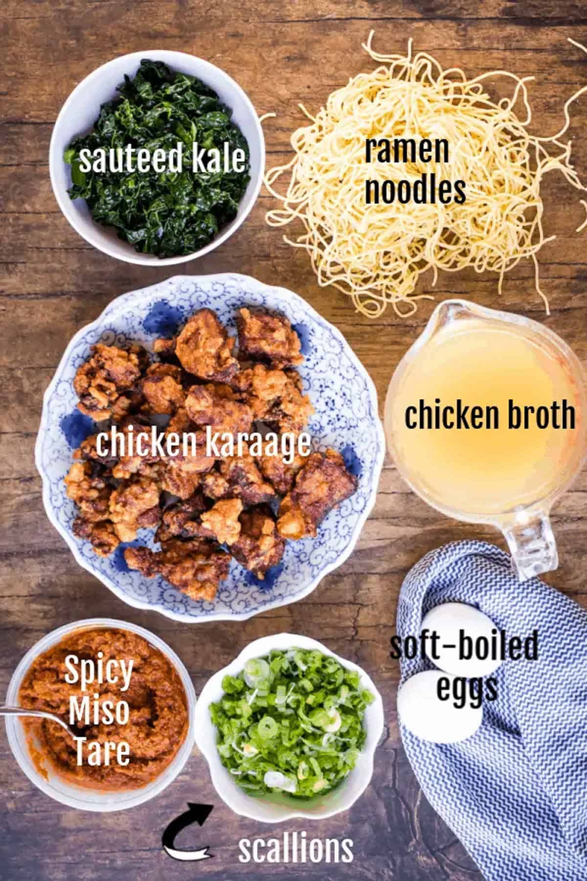 Overhead view of ingredients for miso ramen: sautéed kale, ramen noodles, chicken karaage, chicken broth, spicy miso tare, scallions, and soft-boiled eggs arranged on a wooden table.