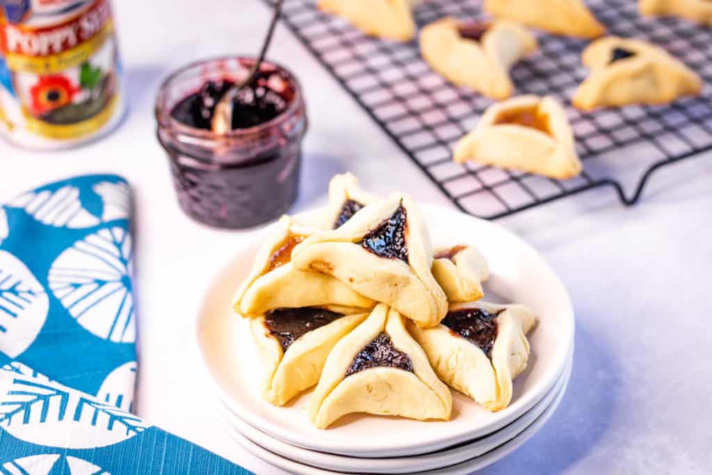 hamentashen cookies piled on a white plate with more cookies on a rack in the background. There is jar of jam with a spoon in it in the background too.