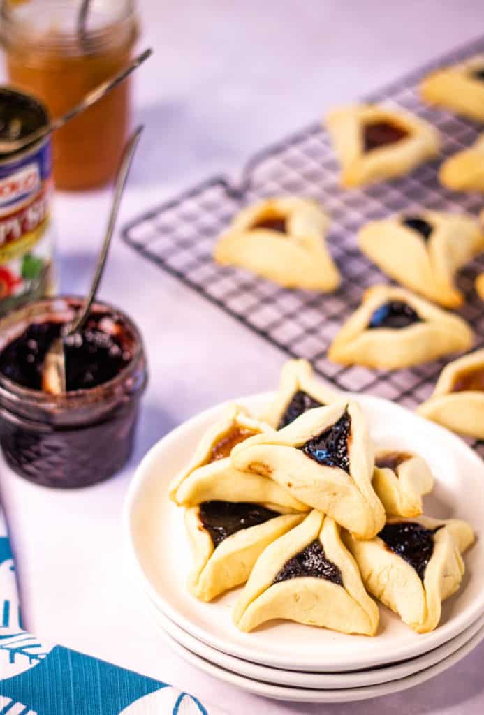 low angle shot of baked hamentashen cookies. There are a bunch of cookies piled up on a white palte, more cookies on a cooling rack in the background.