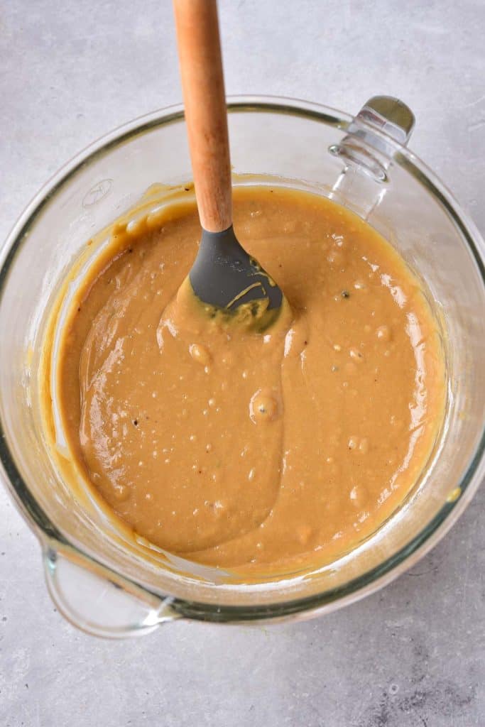 A glass bowl filled with a thick, creamy honey cake batter being stirred with a silicone spatula on a gray surface.
