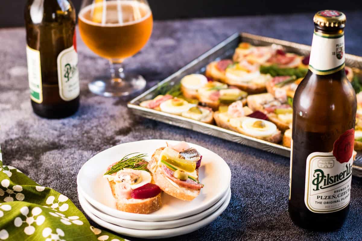 Low angle shot of a stack of white plates with two open face sandwiches on the top plate. There are two bottles of beer, a glass of beer, and a tray of open faced sandwiches in the background. 