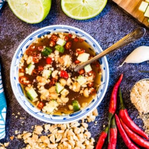 overhead photo of a bowl of thai dipping sauce with limes, chilies, sugar, garlic, crushed peanuts on the table around the bowl.