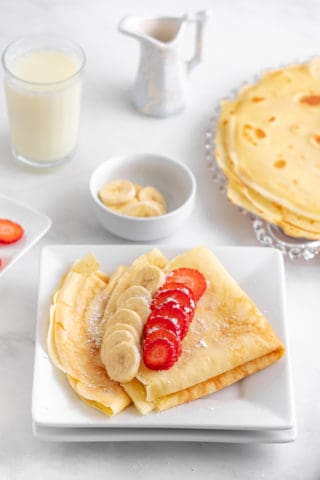 Low angle shot of crepes folded on a square plate with strawberries and bananas on top with a bowl of banana slices and a stack of cooked crepes in the background.