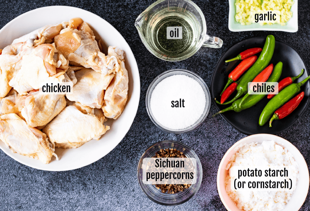 Ingredients for Chinese salt and pepper chicken wings arranged on a countertop: raw chicken pieces, oil, salt, Sichuan peppercorns, potato starch or cornstarch, chiles, and chopped garlic.