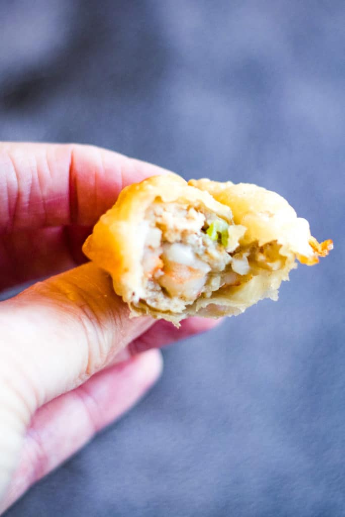 Shot of a hand holding a fried wonton with a bite taken out of it so that you can see the pork and shrimp filling inside