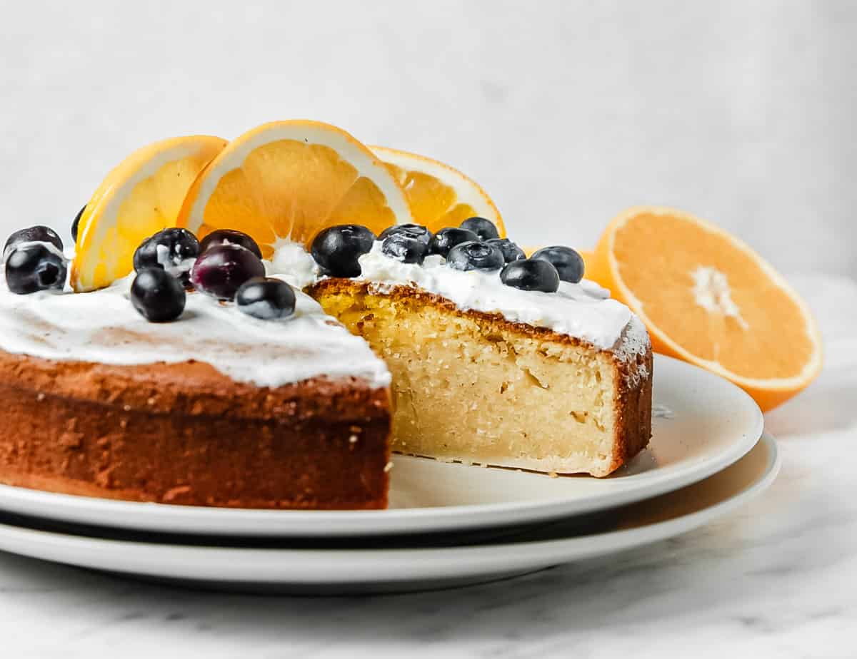 low angle shot of the orange sponge cake with a slice removed. It is topped with whipped cream, blueberries, and orange slices.
