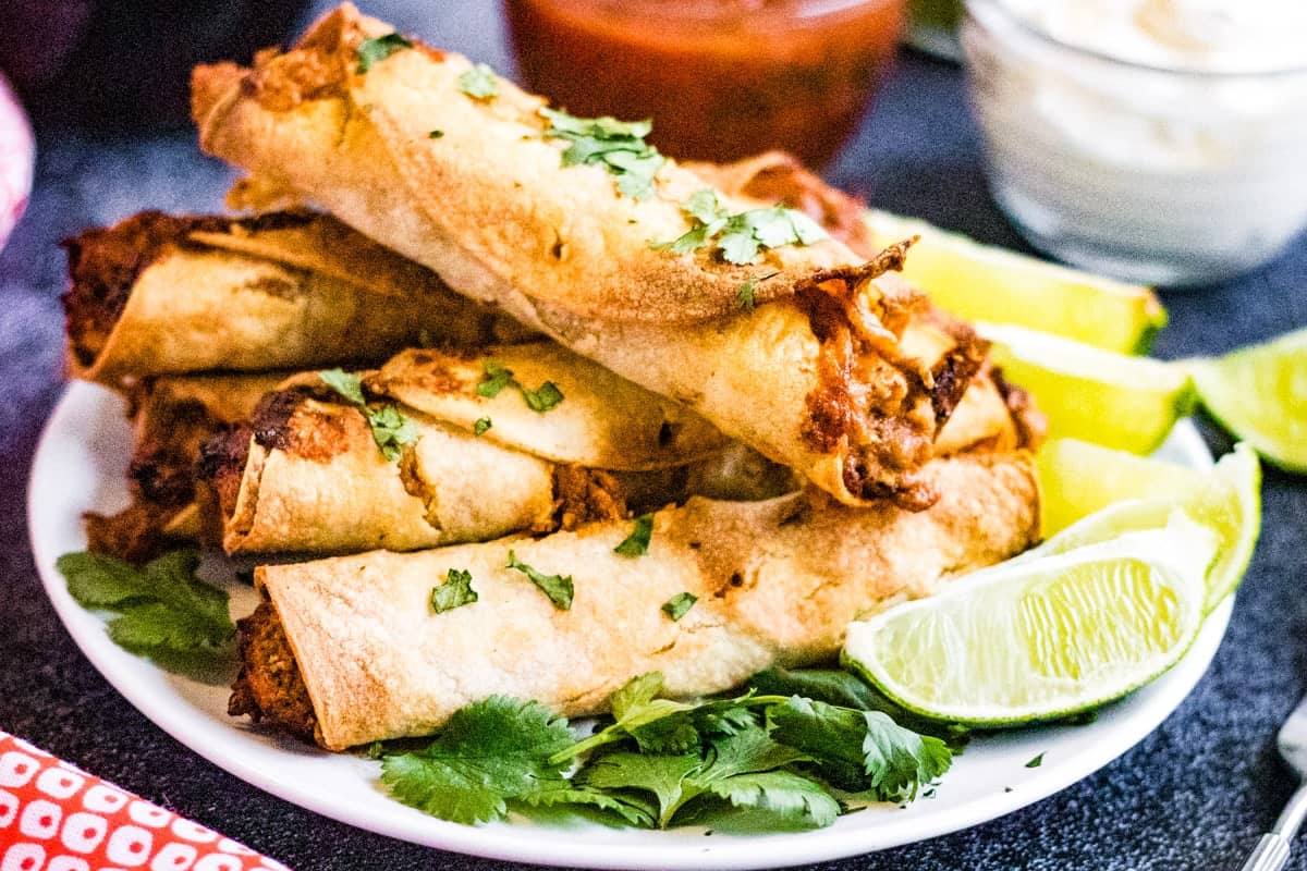 Taquitos piled on a plate. There are lime wedges on the side and they're garnished with chopped cilantro.