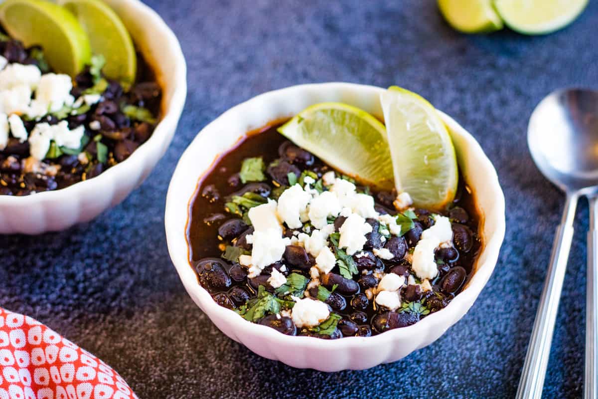 A low-angle shot of two white bowls filled with black beans garnished with cheese, cilantro, and lime slices.