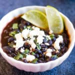 Low angle shot of a white bowl with black breans garnished with cilantro, crumbled feta cheese, and lime wedges.