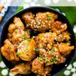 A bowl of air fryer orange chicken garnished with sesame seeds and chopped chives, set on a green and white polka dot background. Text overlay reads "Air Fryer Orange Chicken.