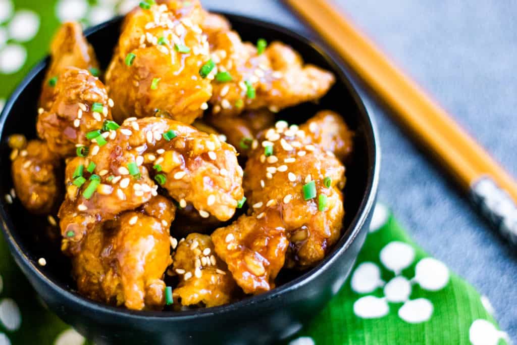 low angle shot of air fryer orange chicken in a black bowl. Garnished with chives and the bowl is sitting on a green and white flowered napkin.