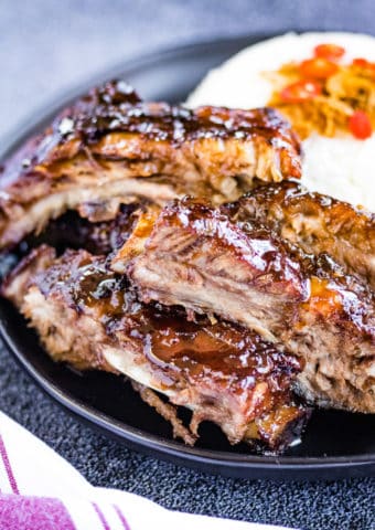 A low angled shot of the cooked ribs on a black plate with rice in the background.