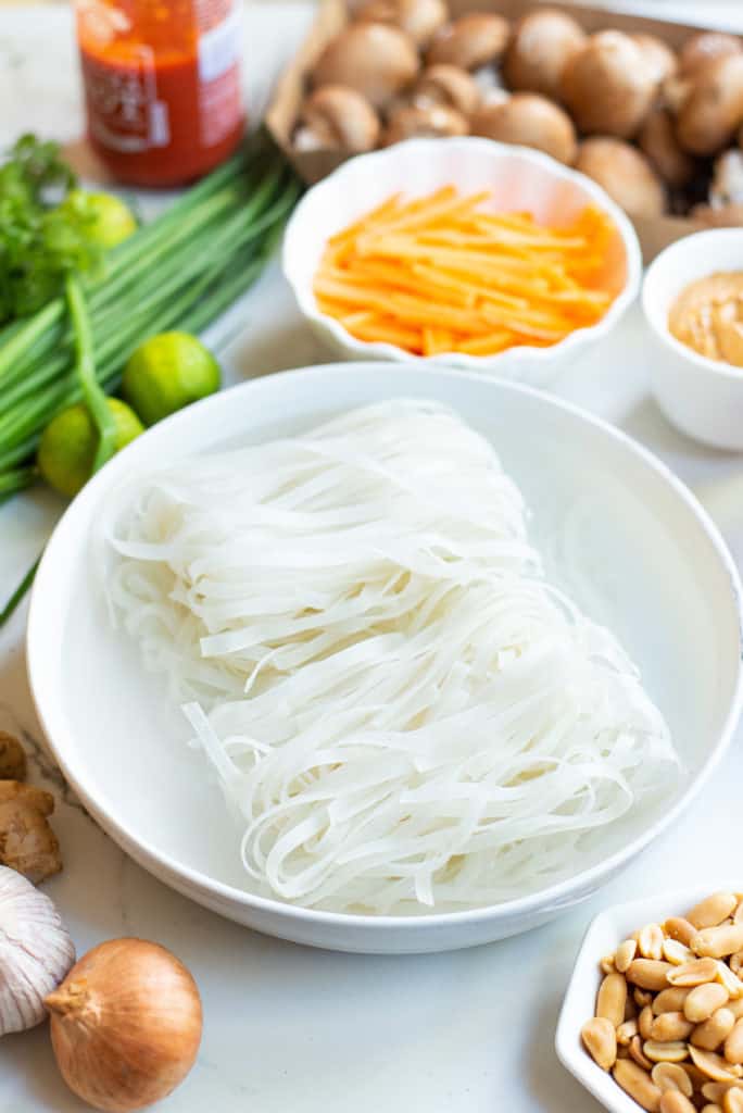 rice noodles soaking in a bowl of water with vegetables and peanuts on the side.