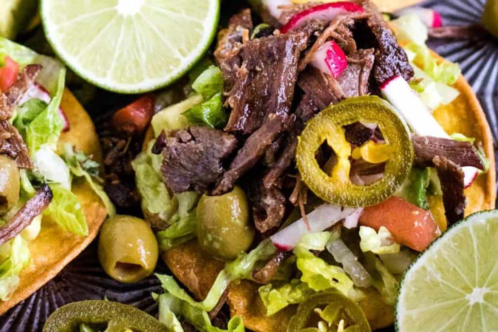 Close-up of a tostada topped with shredded beef salpicon, lettuce, pickled jalapeños, olives, diced vegetables, and garnished with lime slices.