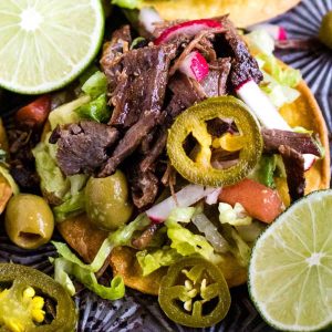 A tostada topped with salpicon—shredded beef, lettuce, radish, tomato, green olives, and sliced jalapeños—surrounded by lime wedges.