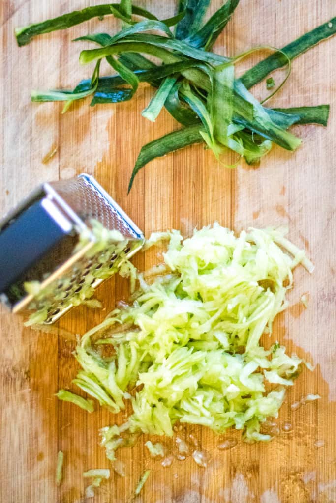 A grater on a cutting board with cucumber raita and some scallions.