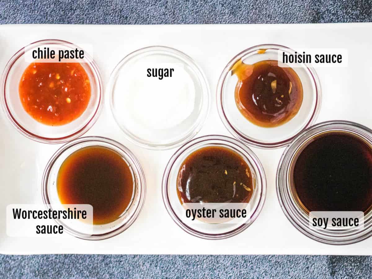 Six small bowls on a white tray contain chile paste, sugar, hoisin sauce, Worcestershire sauce, oyster sauce, and soy sauce—each labeled with text—perfect for customizing dishes like beef yakisoba.