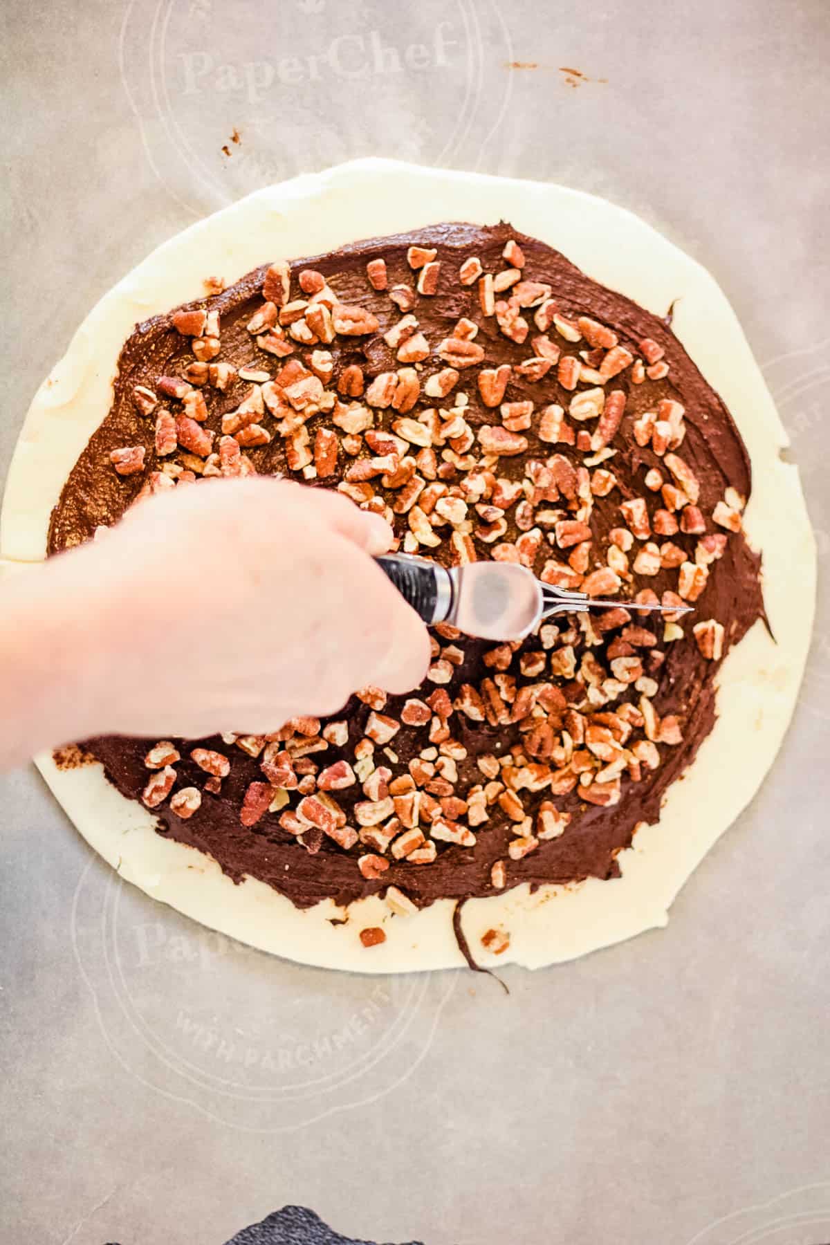 A person incorporating rugelach and pecans into a chocolate pizza creation.
