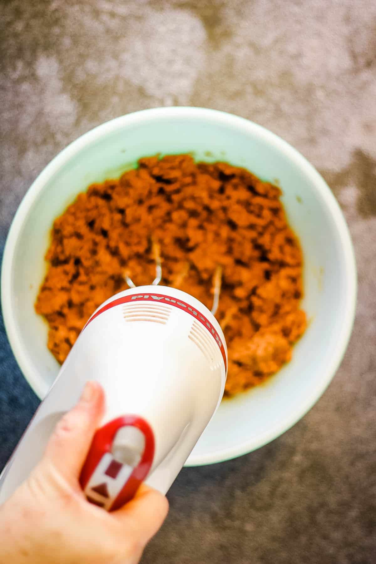 A person using a hand mixer to mix ingredients for ginger snap cookies in a bowl.