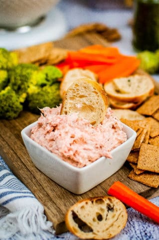A bowl of smoked salmon dip with crackers and broccoli on a cutting board.