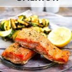 Two pieces of perfectly cooked air fryer salmon sit on a glass plate, surrounded by grilled vegetables and a lemon wedge. "Air Fryer Salmon" is elegantly displayed at the top, with "allwaysdelicious.com" noted at the bottom.