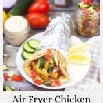 A chicken shawarma wrap in foil on a plate with cucumbers and tomatoes, beside ingredients and a labeled jar, with text "air fryer chicken shawarma.