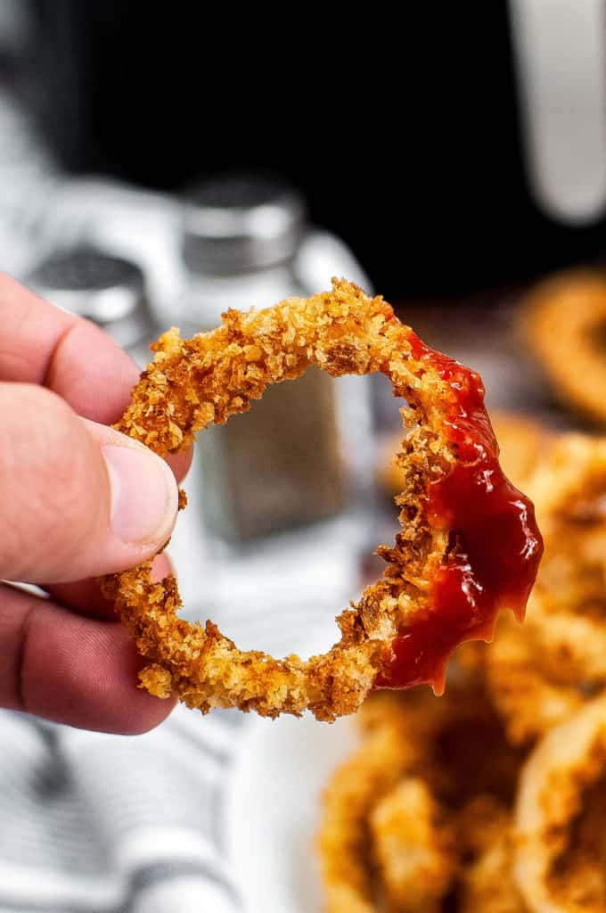 Closeup shot of an onion ring being held up by someone's hand. The onion ring has been dipped in ketchup.