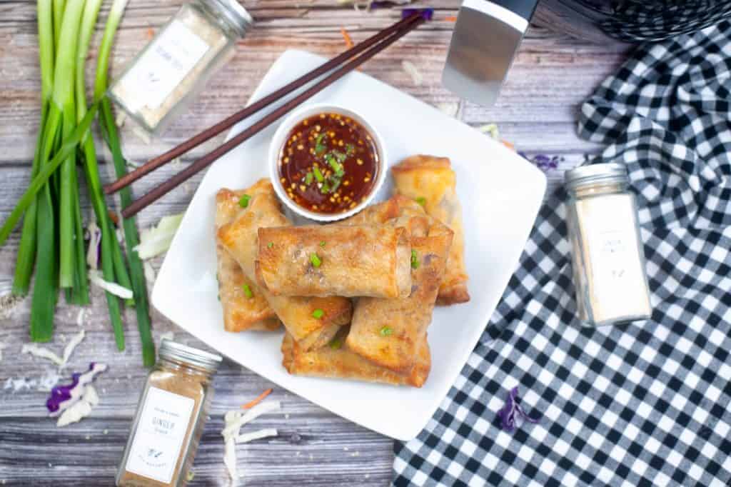A plate of golden-brown air fryer spring rolls with dipping sauce, chopsticks, sliced green onions, spice jars, and a checkered napkin on a wooden table.