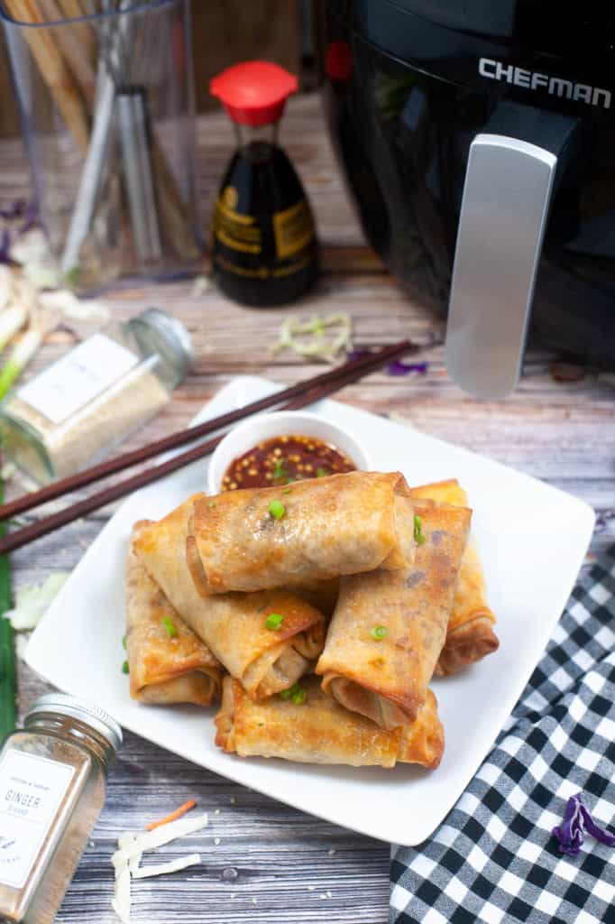 low-angle shot of spring rolls piled on a white plate with the air fryer and a bottle of soy sauce in the background.