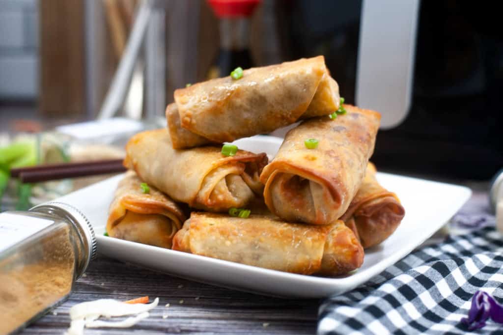 low-angle shot of spring rolls piled on a white plate with the air fryer and a bottle of soy sauce in the background.