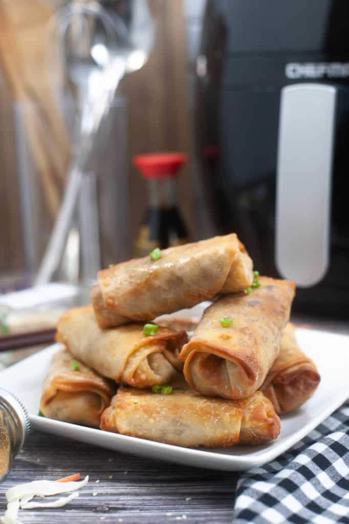 Straight on shot of spring rolls piled on a white plate with the air fryer and a bottle of soy sauce in the background.