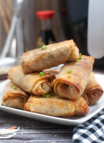 A stack of golden-brown air fryer spring rolls garnished with chopped green onions on a white plate.