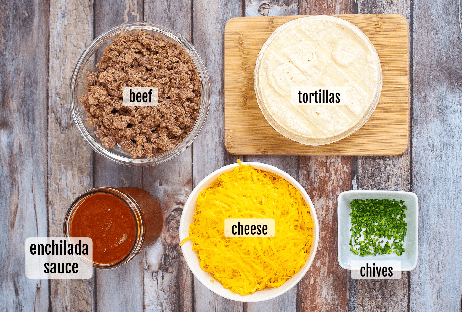 Five ingredients for air fryer enchiladas are displayed: cooked ground beef, corn tortillas, shredded cheese, enchilada sauce in a jar, and chopped chives in a small bowl.
