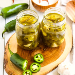 high angle shot of 2 jars of pickled jalapeno peppers with a sliced pepper next to them.