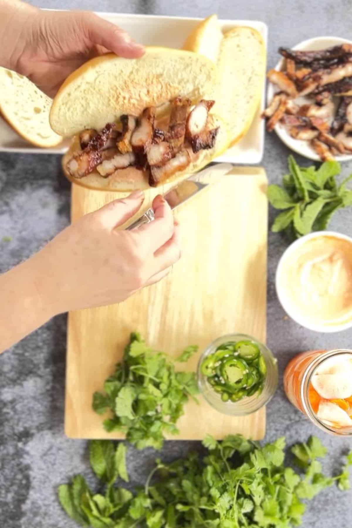 A person is preparing a pork belly banh mi sandwich on a cutting board.