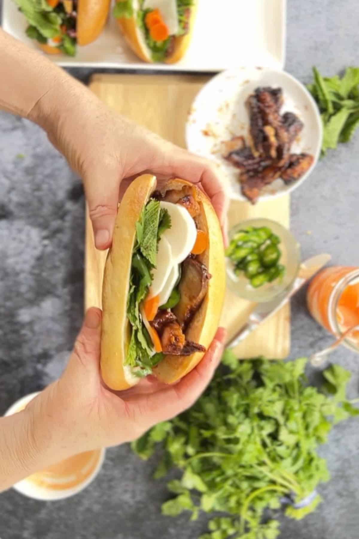 A person holding a pork belly banh mi on a cutting board.