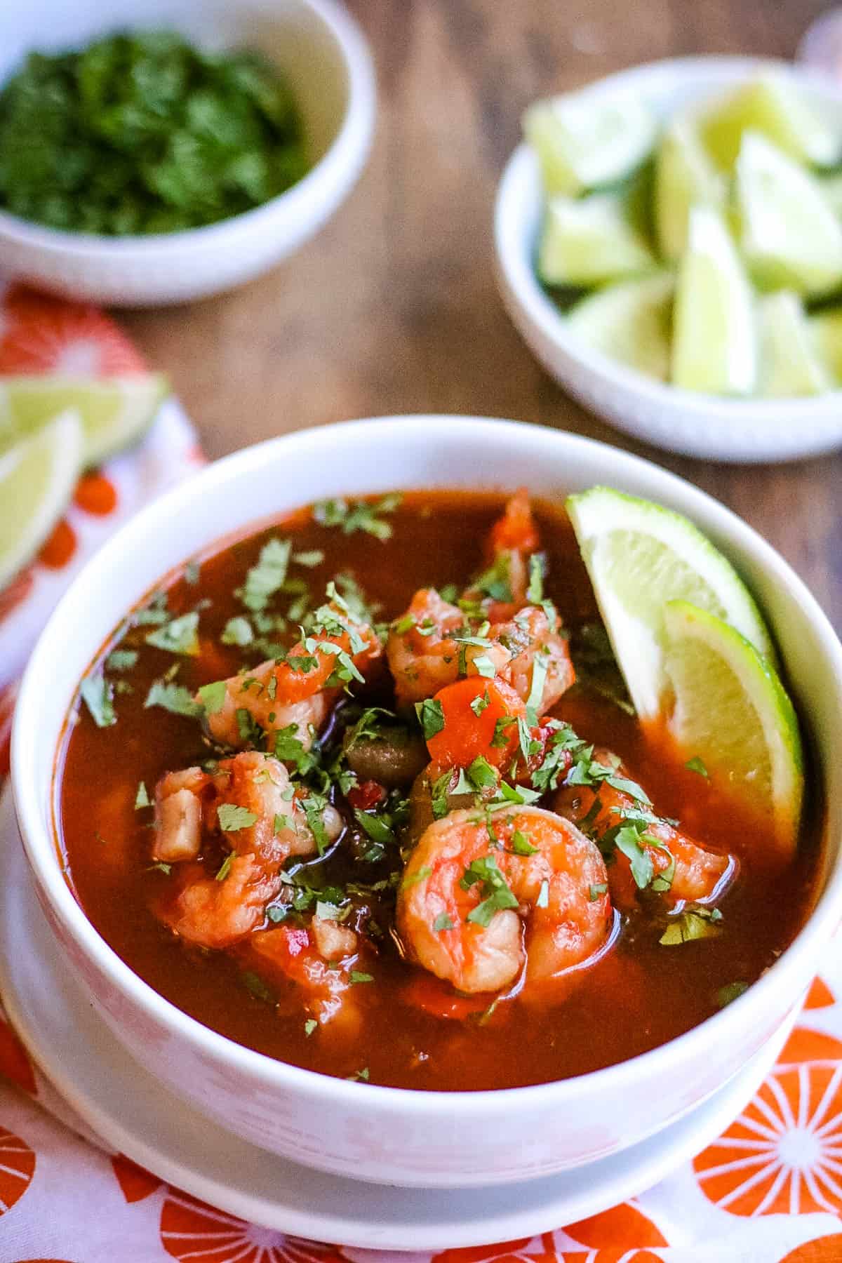 Shot of a bowl of sopa de camarones with lime wedges and chopped cilantro in the background.