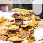 A stack of air fryer s'mores made with graham crackers, melted chocolate, and marshmallows on a plate. A marshmallow and broken pieces of graham crackers are scattered around. Text: "Air Fryer S'Mores - Indulge in the Perfect Blend of Crispy and Gooey!