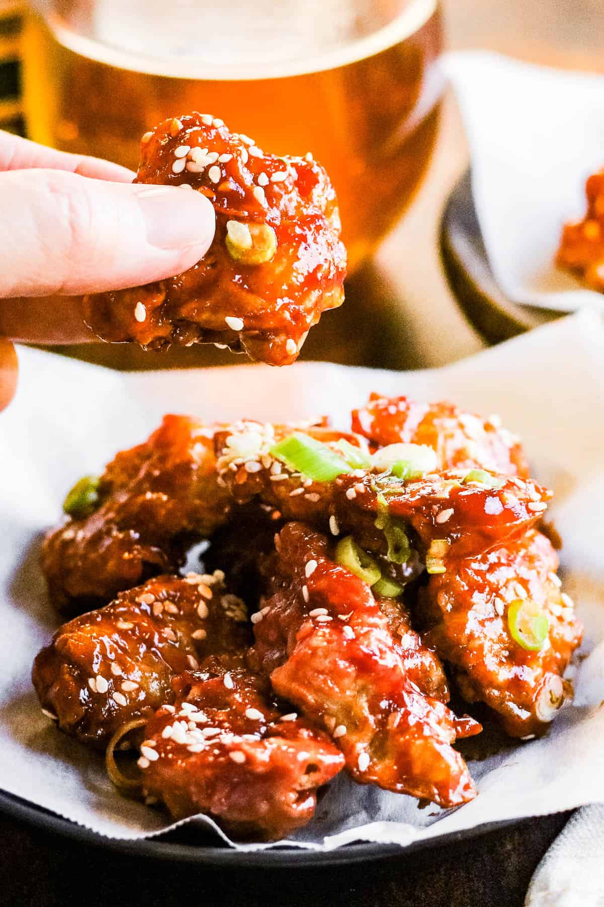Korean fried chicken on a plate with a beer and someone holding a piece of the chicken in their fingers.